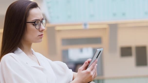 Jovem mulher em roupas de negócios elegantes e óculos usa um smartphone para se comunicar com um chatbot . — Vídeo de Stock