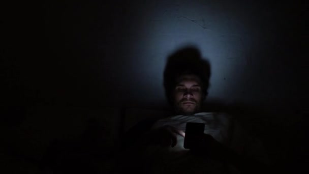 Молодий чоловік в ліжку вночі використовує смартфон. Безсоння і цифрова залежність, дівчина в ліжку — стокове відео