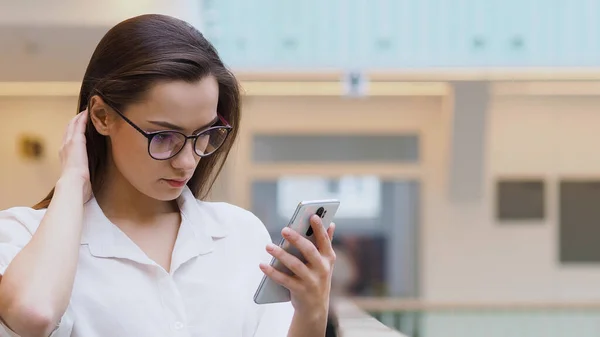 Młoda kobieta w białej koszuli i okularach używa smartfona do wyszukiwania informacji w Internecie. — Zdjęcie stockowe