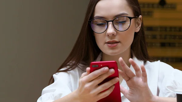 Jovem com óculos usa um smartphone, close-up. O estudante segura em suas mãos o telefone vermelho — Fotografia de Stock