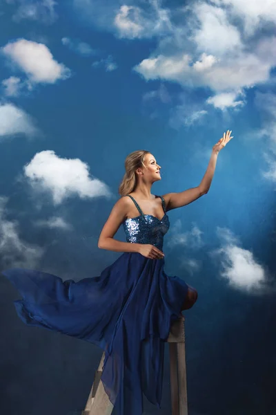 Mädchen auf der Leiter greift mit den Händen nach oben. Eine junge Frau in einem blauen Kleid auf blauem Hintergrund — Stockfoto