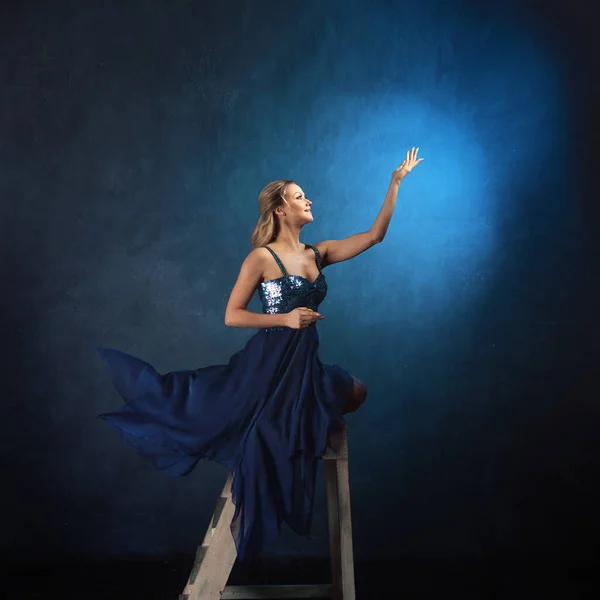 Chica en la escalera alcanza con las manos. Una joven con un vestido azul sobre un fondo azul —  Fotos de Stock