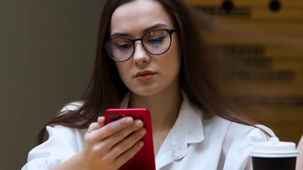 Chica joven con gafas utiliza un teléfono inteligente, primer plano. El estudiante sostiene en sus manos el teléfono rojo — Vídeos de Stock