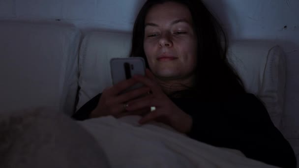 Mujer joven en la cama por la noche utiliza un teléfono inteligente. Insomnio y dependencia digital, la chica en la cama — Vídeos de Stock