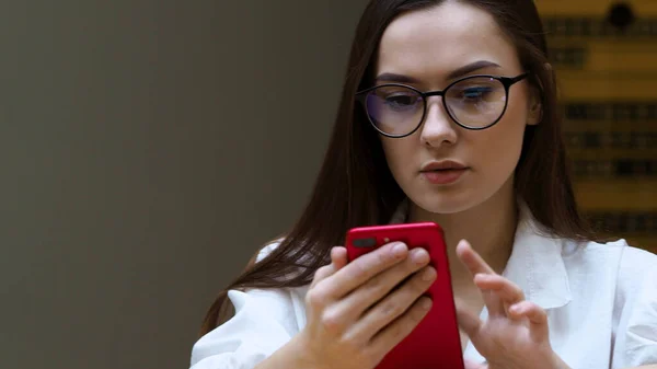 Jovem com óculos usa um smartphone, close-up. O estudante segura em suas mãos o telefone vermelho — Fotografia de Stock