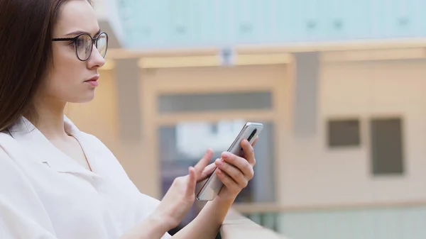Jovem mulher em uma camisa branca e óculos usa smartphone para procurar informações na Internet . — Fotografia de Stock