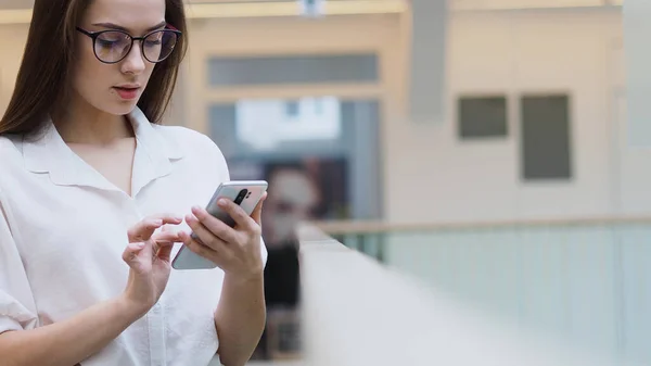 Jovem mulher em uma camisa branca e óculos usa smartphone para procurar informações na Internet . — Fotografia de Stock