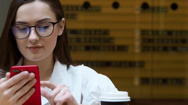 Fiatal lány szemüveggel okostelefont használ, közelről. A diák kezében tartja a piros telefont. — Stock Fotó