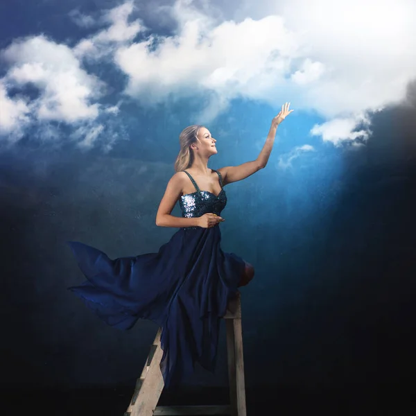 Mädchen auf der Leiter greift mit den Händen nach oben. Eine junge Frau in einem blauen Kleid auf blauem Hintergrund — Stockfoto