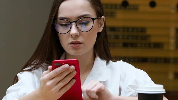 Jong meisje met een bril gebruikt een smartphone, van dichtbij. De student houdt de rode telefoon in zijn handen — Stockfoto