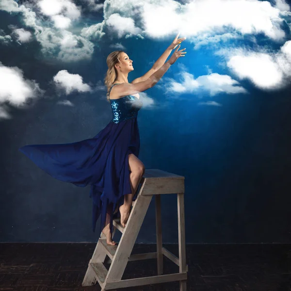 Chica en la escalera alcanza con las manos. Una joven con un vestido azul sobre un fondo azul —  Fotos de Stock