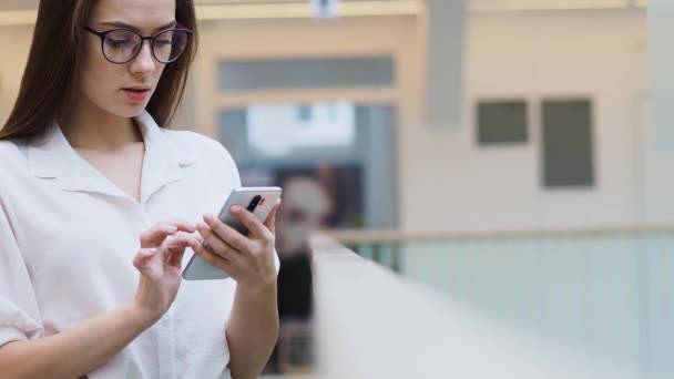 Mujer joven con una camisa blanca y gafas utiliza el teléfono inteligente para buscar información en Internet . — Vídeo de stock