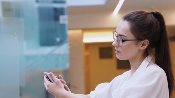 Joven dama de negocios utiliza un teléfono inteligente. Una chica con una camisa blanca y gafas hace una llamada . — Vídeo de stock