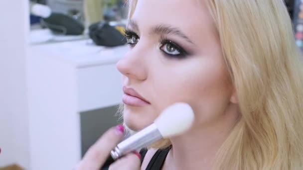 Jonge blonde vrouw in een sessie met een make-up kunstenaar, make-up kunstenaar brengt poeder, — Stockvideo