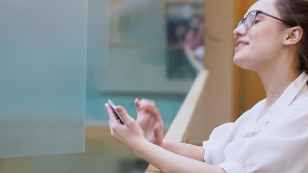 Jovem empresária usa um smartphone. menina surpreso na camisa branca e óculos faz um telefonema . — Vídeo de Stock