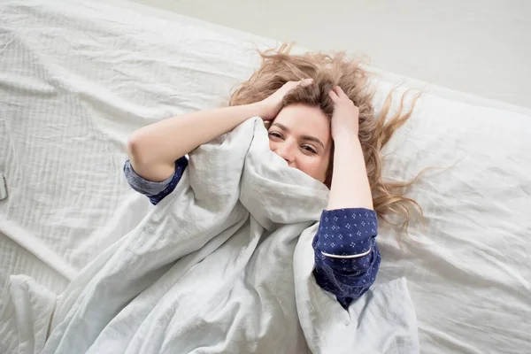 Linda rubia en su cama en pijama azul, debajo de una manta — Foto de Stock