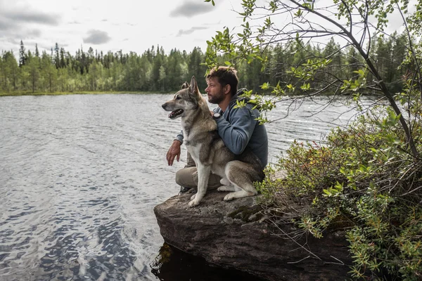 Handsome man and dog are sitting on the stone near of lake and looking into the distance — Stock Photo, Image