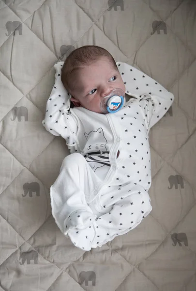 Pensive new born baby boy is laying in white sleepers with blue nipple and watching into the window — Stock Photo, Image