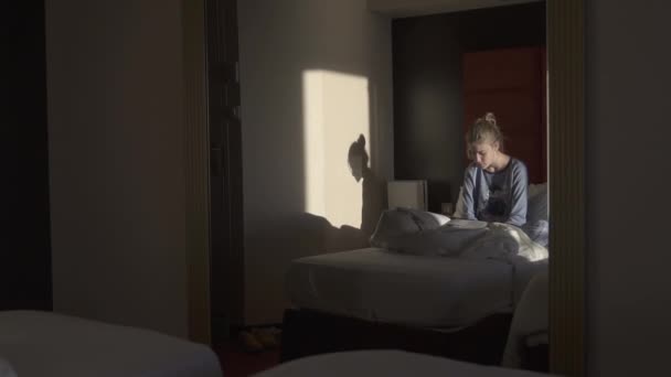 Piękna dziewczyna leży w łóżku i czyta książkę w domu lub hotelu — Wideo stockowe