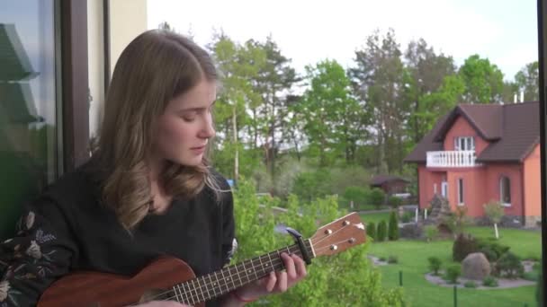 Vacker Tonåring Flicka Spelar Ukulele Gitarr Sitter Fönstret Tröskel Bakgrunden — Stockvideo