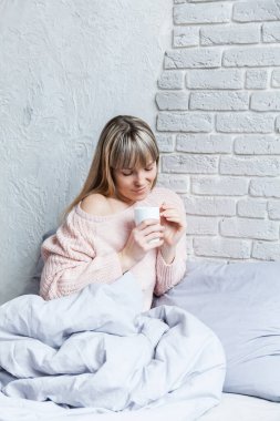 Genç bir kadın yatakta otururken kahvesini içiyor. Yatakta güneşli bir sabah.