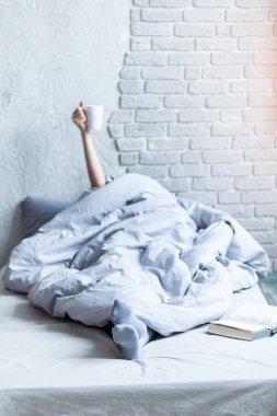 Kadın battaniyenin altında, kollarını kaldırmış, elinde bir fincan kahve tutuyor ve zafer işareti gösteriyor.