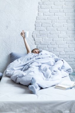 Kadın battaniyenin altında, kollarını kaldırmış, elinde bir fincan kahve tutuyor ve zafer işareti gösteriyor.