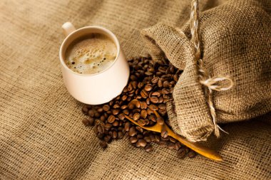 Bir fincan aromatik kahve, tarçın çubukları, dağınık kahve taneleri bir poşetten ve çuval bezi kumaşında ahşap kepçe