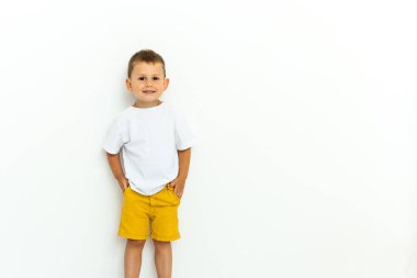 Sarı şortlu, beyaz tişörtlü, beyaz arka planda elleri ceplerinde sevimli bir çocuk.
