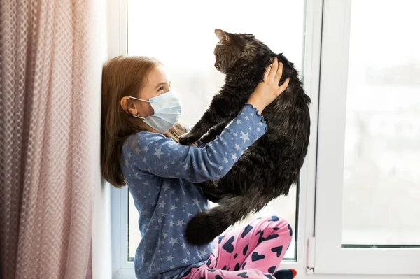 Girl in medical mask holding cat and suffering from allergy at home on the window. COVID-19. Pandemic Coronavirus. Kid home quarantine wearing face mask protective
