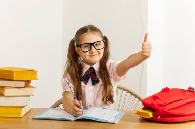 Masasında açık not defteri ve okul çantasıyla gözlüklü mutlu küçük bir kız, baş parmak işareti gösteriyor. Eğitim kavramı.