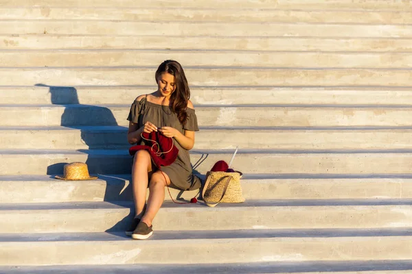 A young woman sitting In a deserted place cityscape, on the steps and knitting a sweater with knitting needles on a summer day. Get Creative everywhere during quarantine