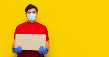 Tıbbi maskeli bir teslimatçı sarı arka planda izole edilmiş bir karton kutu taşıyor. Karantina konseptiyle internetten yiyecek al. Pankart