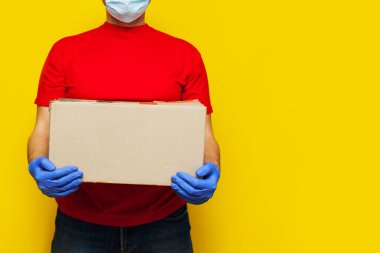 Tıbbi maskeli bir teslimatçı sarı arka planda izole edilmiş bir karton kutu taşıyor. İnternetten karantina konsepti satın al.