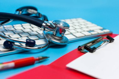 Bilgisayar klavyesinde steteskop, mavi arka planda kırmızı pano ve kalem. Tıbbi bakım konsepti, üst görünüm, düz alan, kopyalama alanı