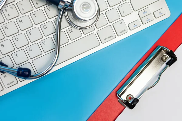 Stethoscope on computer keyboard and red Clipboard on Blue background. Medical care treatment concept, top view, flat lay, copy space