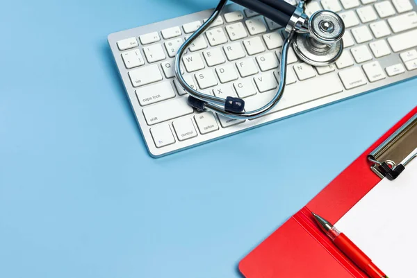 Stethoscope on computer keyboard and red Clipboard and pen on Blue background. Medical care treatment concept, top view, flat lay, copy space