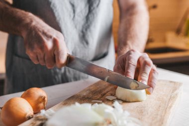 Kaba erkek elleri mutfağın arka planında beyaz bir soğanı bıçakla kesiyor.
