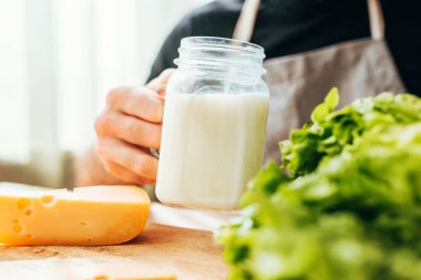 Pencere kenarındaki masada oturan bir adam süt içer ve peynirli ve otlu kahvaltı yapar.