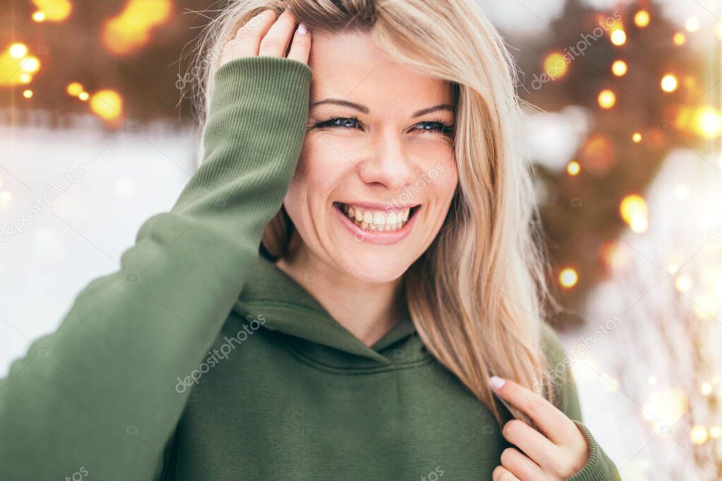 Outdoor portrait of an attractive cute blonde with a wide smile, white teeth and big eyes - against the background of lights - wearing a hoodie
