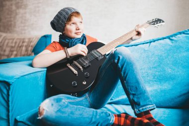 Kot pantolonlu, şapkalı ve kırmızı tişörtlü çekici çocuk evdeki kanepede oturan siyah elektro gitarlı bir gülümsemeyle rüya gibi oynuyor.