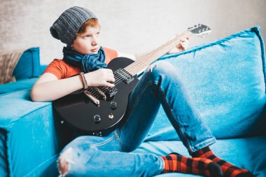 Kotlu, şapkalı ve kırmızı tişörtlü çekici çocuk evdeki kanepede oturan siyah bir elektro gitarda çalıyor.