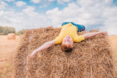 İngiliz kızıl saçlı, neşeli çocuk baş aşağı bir tarlada istiflenmiş bir şekilde uzanıyor ve parlak güneşe bakıyor.