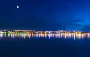 Deniz kıyısının inanılmaz renkli gece fotosu. Şehir ışıklarının sudaki yansıması.