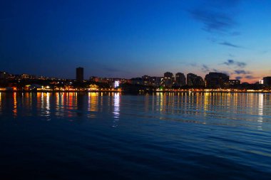 Deniz kıyısında inanılmaz renkli bir gün batımı. Şehir ışıklarının sudaki yansıması.