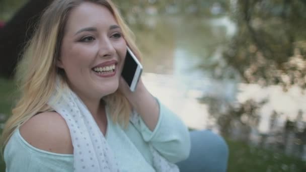 Mujer rubia más tamaño hablar Smartphone sentarse junto al lago — Vídeo de stock