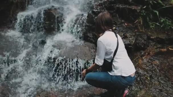 Female hiker crouch at splashing jungle waterfall — Stock Video