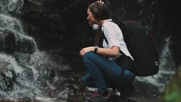 女旅行家蹲伏在飞溅的瀑布边 — 图库视频影像