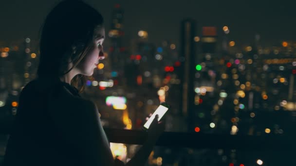 Όμορφη κοπέλα περιηγηθείτε κινητό τηλέφωνο νύχτα οροφή της πόλης — Αρχείο Βίντεο