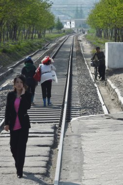 Kaesong, North Korea - May 5, 2019:  Local people go down along railway in industrial district of  Kaesong, one of the North Korean city. Selective focus on the women with red flag clipart
